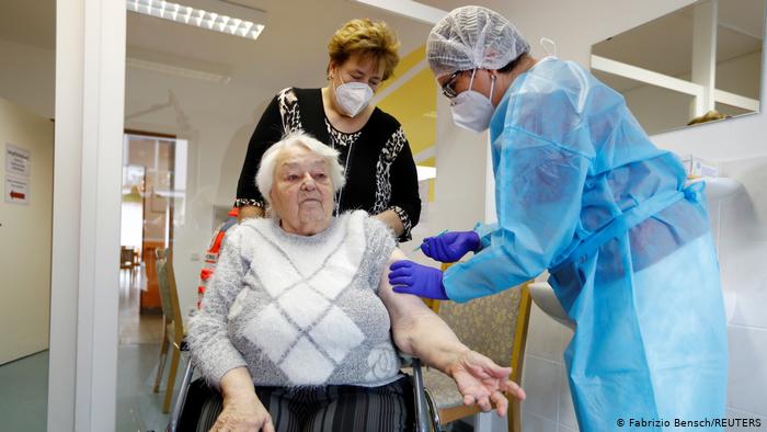 Ruth Heise sitting in a wheelchair, receiving an injection from a doctor in protective gear, as the head of the nursing home looks on